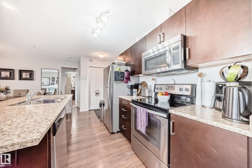 Contemporary kitchen featuring wood-finish flooring, granite-style countertops, stainless steel appliances, dark wood cabinetry, and an undermount sink - 311 142 Ebbers Boulevard, Edmonton, AB 