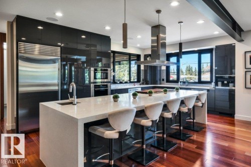 Gourmet kitchen featuring a large waterfall island with integrated sink, stainless steel appliances, dark high-gloss cabinetry, and rich wood-finish flooring - 8914 120 Street, Edmonton, AB 