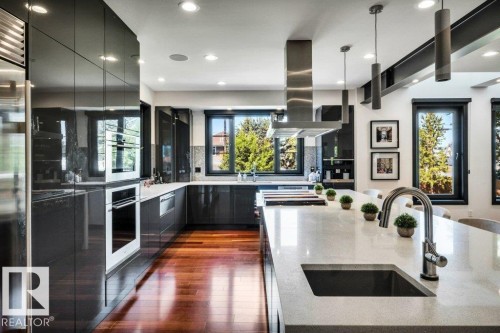 Contemporary kitchen featuring sleek dark cabinetry, wood-finish flooring, a stainless steel range hood, recessed lighting, and a kitchen island with an integrated sink and gooseneck faucet - 8914 120 Street, Edmonton, AB 
