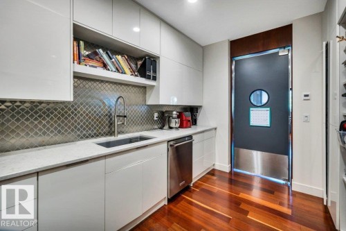 Kitchenette featuring glossy white cabinetry, a stainless steel sink with a modern faucet, light-toned countertops, and a patterned metal tile backsplash - 8914 120 Street, Edmonton, AB 