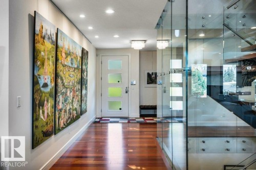 Contemporary entry hallway featuring rich wood-finish flooring, recessed ceiling lighting, a modern white entry door with frosted panels, and a glass-paneled staircase with dark wood treads - 8914 120 Street, Edmonton, AB 