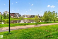 Lakeside setting featuring a paved pathway, expansive green space, mature trees, and residential properties in the background - 