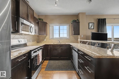 U-shaped kitchen featuring dark wood cabinetry, light granite countertops, stainless steel appliances, a tiled backsplash, and wood-finish flooring - 415 263 Macewan Road, Edmonton, AB 