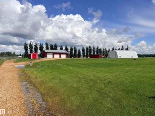 Expansive green lawn featuring multiple outbuildings, including a three-bay red barn with white garage doors, a smaller red shed, and a large white arched structure - 26418 Twp Rd 633, Rural Westlock County, AB 
