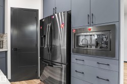 Modern kitchen area featuring a stainless steel refrigerator, built-in microwave, grey cabinetry with dark hardware, and a dark paneled door - 