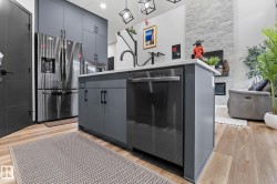 Contemporary kitchen featuring a central island with integrated dishwasher, stainless steel appliances, abundant cabinetry, wood-finish flooring, and a stone accent wall - 