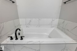 Built-in drop-in bathtub featuring a deck-mounted matte black faucet, surrounded by marbled-patterned tile, and framed by light gray walls - 