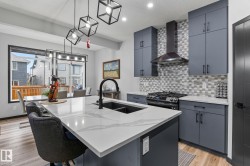 Modern kitchen featuring a large island with white-veined countertop and integrated sink, gray cabinetry with black hardware, a gas range with a black range hood, and a geometric tile backsplash - 