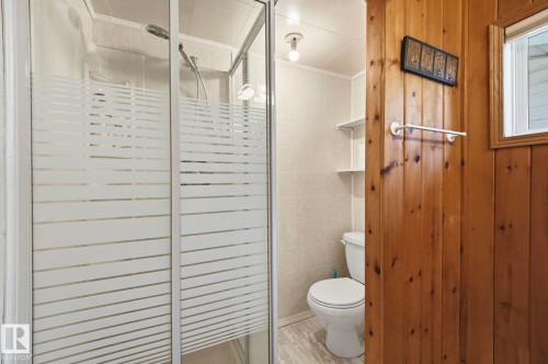 Shower enclosure with frosted glass doors, white ceramic tile wall, light-toned wood paneling, white toilet, and gray-toned floor tile - 1410 47436 Range Rd. 15, Rural Leduc County, AB 