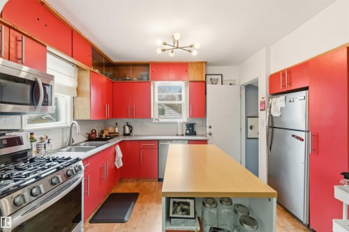 Kitchen featuring red cabinetry, stainless steel appliances including a gas range and microwave, a double basin sink, and wood-finish flooring - 6036 106 Street, Edmonton, AB 