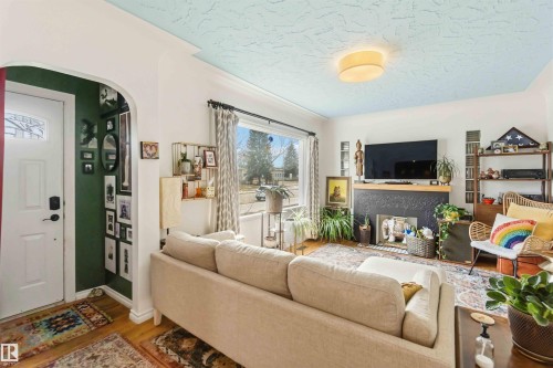 Living area featuring wood-finish flooring, a decorative fireplace with a dark-toned surround, a front entry door with a decorative glass insert, and a large window - 6036 106 Street, Edmonton, AB 