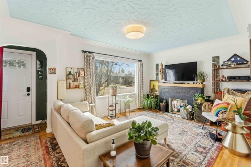 Living area featuring a textured light blue ceiling, wood-finish flooring, a prominent picture window, and a fireplace with a dark-finished mantel - 6036 106 Street, Edmonton, AB 