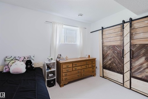 Room featuring barn-door style closet doors with herringbone pattern, white walls, light-colored carpeting, and a window with white drapes - 1743 Melrose Crescent Sw, Edmonton, AB 
