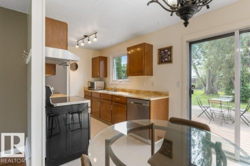 Kitchen featuring wood cabinetry, light-toned countertops, a stainless steel dishwasher, and a black range with stainless steel hood - 18609 54 Avenue, Edmonton, AB 