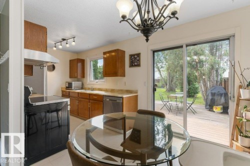 Kitchen dining area featuring a glass-top table, sliding patio doors, a wooden deck, and a fenced yard with mature trees - 18609 54 Avenue, Edmonton, AB 