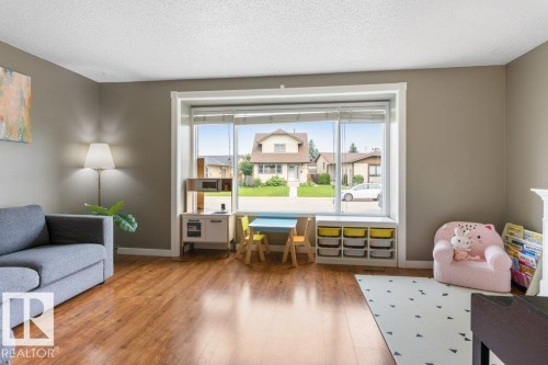 Spacious living area featuring expansive front-facing window, wood-finish flooring, neutral wall tones, and white baseboards - 18609 54 Avenue, Edmonton, AB 
