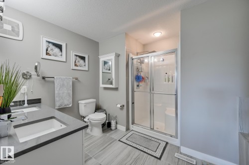 Bathroom featuring a dual vanity with undermount sinks and dark countertops, a shower with sliding glass doors, a toilet, and light gray wood-finish tile flooring - 101 Hilldowns Drive, Spruce Grove, AB 