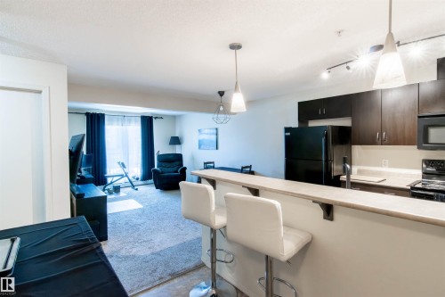 Open-concept living space featuring a kitchen with dark cabinetry, light countertops, and black appliances - #214 1188 Hyndman Rd Nw, Edmonton, AB - Indoor Photo Showing Kitchen