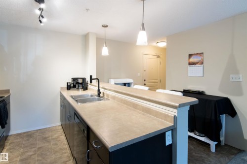 Contemporary kitchen featuring dark cabinetry, light countertops, and a double basin stainless steel sink with a matte black faucet - #214 1188 Hyndman Rd Nw, Edmonton, AB - Indoor Photo Showing Kitchen With Double Sink