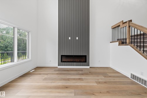 Vaulted ceiling living space featuring a floor-to-ceiling accent wall with a linear electric fireplace, large windows, wood-finish flooring, and a staircase with wood handrails and black metal balusters - 915 Elderberry Landing Nw, Edmonton, AB - Indoor Photo Showing Other Room With Fireplace