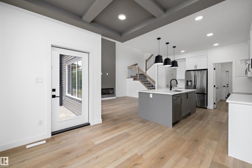 Open-concept living space featuring wood-finish flooring, a modern kitchen island with pendant lighting, stainless steel appliances, a contemporary fireplace, and a striking coffered ceiling - 915 Elderberry Landing Nw, Edmonton, AB - Indoor Photo Showing Kitchen With Upgraded Kitchen