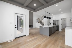 Open-concept living space featuring wood-finish flooring, a modern kitchen island with pendant lighting, stainless steel appliances, a contemporary fireplace, and a striking coffered ceiling - 