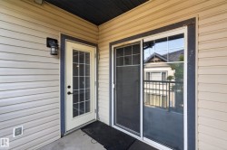 Outdoor balcony featuring a white-framed glass paneled door, a sliding glass door, light beige siding, an exterior wall sconce, and a dark soffit - 