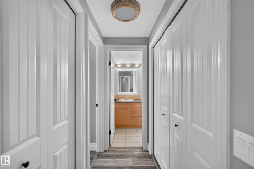 Hallway featuring wood-finish flooring and multiple white bi-fold doors, leading to a room with a wood vanity, light-colored countertop, and wall-mounted light fixture - 18 2565 Hanna Crescent, Edmonton, AB 