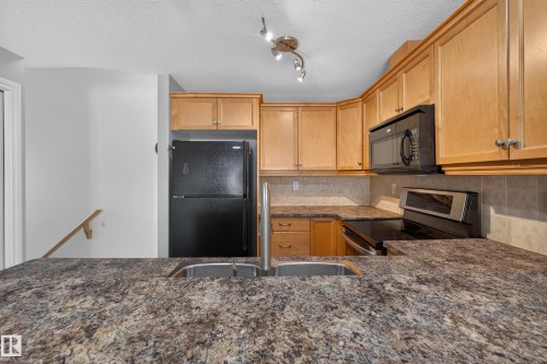 Kitchen featuring a double basin stainless steel sink, granite-finish countertops, wood cabinetry, a black over-the-range microwave, and a black refrigerator - 18 2565 Hanna Crescent, Edmonton, AB 