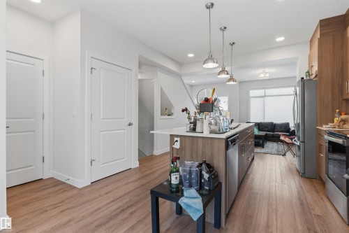 Open-concept kitchen featuring a large island with white countertop, wood-finish flooring throughout, stainless steel appliances, and pendant lighting - 170 804 Welsh Drive, Edmonton, AB 