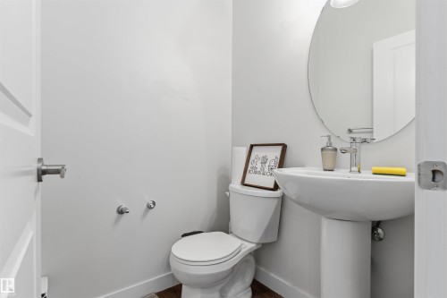 Contemporary powder room featuring a pedestal sink with chrome faucet, a white toilet, a round wall-mounted mirror, and wood-finish flooring - 170 804 Welsh Drive, Edmonton, AB 