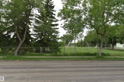 Expansive green space featuring a chain-link fence, mature trees, and a paved sidewalk along a street - 