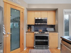 Kitchen area featuring a light wood-finish pantry door with a frosted glass insert, light wood-finish cabinetry, stainless steel appliances, and a dark tile backsplash - 
