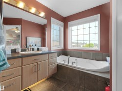 Spacious bathroom featuring an integrated soaking tub with tiled surround, dual windows with white blinds, and a vanity with an integrated sink and light wood-finish cabinetry - 