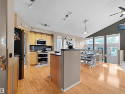 Open-concept kitchen and dining area featuring light wood-finish flooring, stainless steel appliances, a kitchen island with a dark countertop, a vaulted ceiling, and multiple windows - 