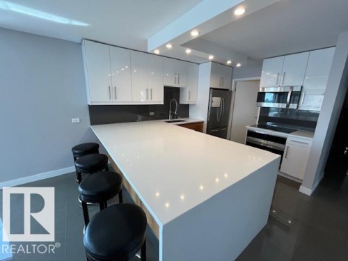 Spacious kitchen island with a waterfall countertop, complemented by sleek white cabinetry, a dark backsplash, stainless steel appliances, and recessed lighting - 1101 10011 123 Street, Edmonton, AB 