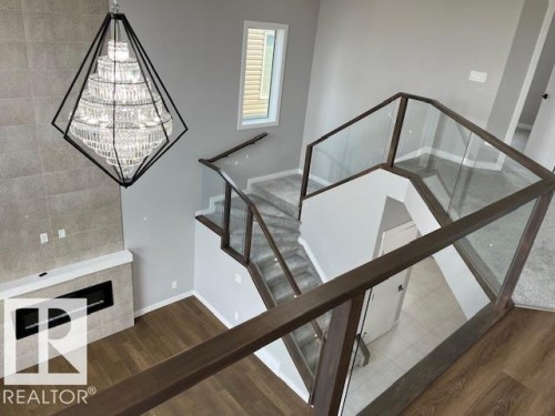 Dramatic two-story entry with a geometric crystal chandelier, contemporary electric fireplace with a tiled surround, wood-finish flooring, and glass-paneled railings with wood-finish handrails - 3810 46 Avenue, Beaumont, AB 