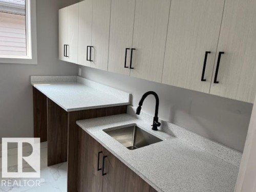 Utility room featuring white shaker-style upper cabinetry with black hardware, wood-finish base cabinetry, light speckled countertops, and a stainless steel sink with a matte black faucet - 3810 46 Avenue, Beaumont, AB 