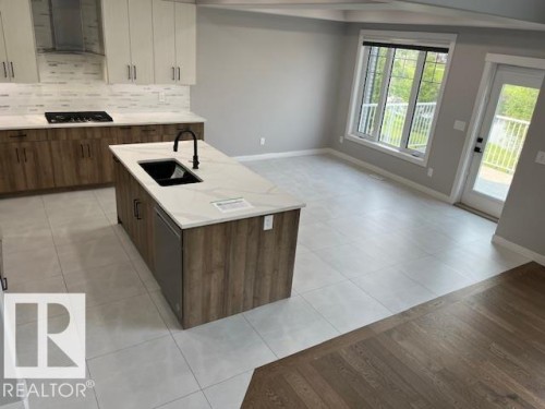 Open-concept kitchen and living area featuring a large center island with integrated sink, wood-finish cabinetry, tile flooring, and a large window with exterior access - 3810 46 Avenue, Beaumont, AB 