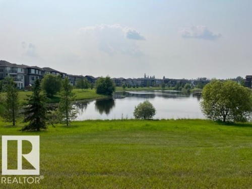 Expansive greenspace bordering a calm body of water, featuring a distant row of multi-story residential structures with balconies - 3810 46 Avenue, Beaumont, AB 