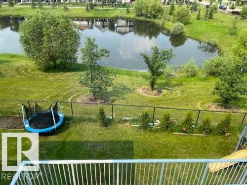 Landscaped backyard featuring a pond with surrounding greenery, a black chain-link fence, and a white balcony railing - 3810 46 Avenue, Beaumont, AB 