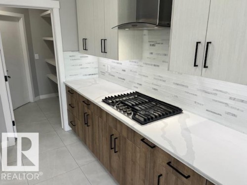 Modern kitchen featuring a gas range with a stainless steel vent hood, white countertops, two-tone cabinetry with matte black hardware, a mosaic tile backsplash, and light-toned floor tile - 3810 46 Avenue, Beaumont, AB 