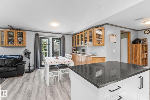 Open-concept living area featuring light wood-finish flooring, a kitchen island with a dark speckled countertop and white base cabinetry, light wood-toned upper and lower cabinetry with glass panel doors, and double French doors with interior window panes - 31 Graywood Cove, Stony Plain, AB 