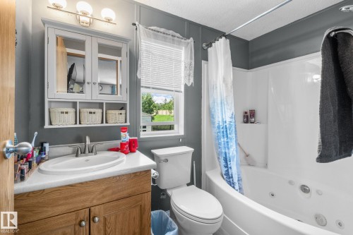Bathroom featuring a jetted tub, wood-finish vanity with an integrated sink, wall-mounted medicine cabinet with open shelving, and a window with blinds - 31 Graywood Cove, Stony Plain, AB 