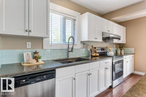 Bright kitchen featuring white cabinetry, dark stone countertops, a light blue subway tile backsplash, a double basin sink with a gooseneck faucet, and stainless steel appliances - 1119 Potter Greens, Edmonton, AB - Indoor Photo Showing Kitchen With Double Sink With Upgraded Kitchen