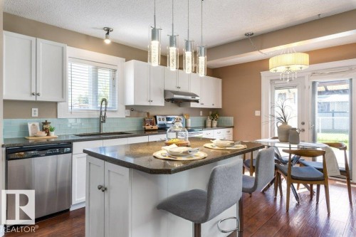 Modern kitchen featuring white cabinetry, a dark countertop island with seating, stainless steel appliances, a light blue subway tile backsplash, and wood-finish flooring - 1119 Potter Greens, Edmonton, AB - Indoor Photo Showing Other Room