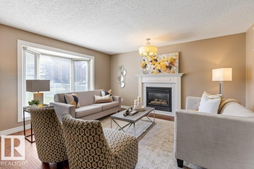 Living area featuring a fireplace with a white mantel, a bay window with blinds, wood-finish flooring, and a central chandelier - 1119 Potter Greens, Edmonton, AB - Indoor Photo Showing Living Room With Fireplace