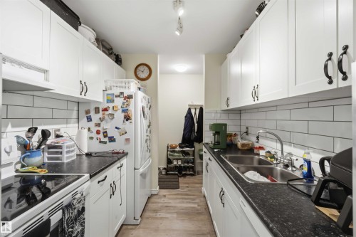 Galley kitchen featuring white cabinetry with black hardware, dark countertops, a subway tile backsplash, and wood-finish flooring - 204 4601 131 Avenue, Edmonton, AB 