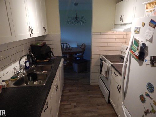 Galley kitchen featuring white cabinetry, dark countertops, a subway tile backsplash, stainless steel sink, and wood-finish flooring - 204 4601 131 Avenue, Edmonton, AB 