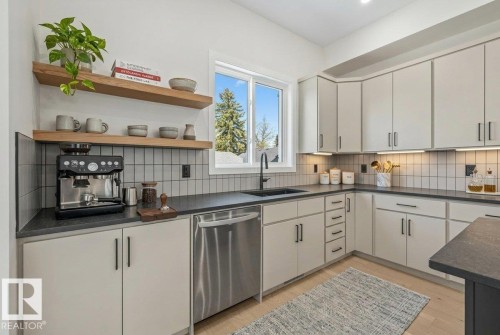 Modern kitchen featuring light cabinetry, dark countertops, a stainless steel dishwasher, a black undermount sink with a matching faucet, and light wood-finish flooring - 10932 132 Street, Edmonton, AB - Indoor Photo Showing Kitchen